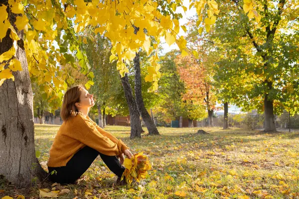 Parlak, güneşli bir günde, turuncu kazaklı 35-40 yaşlarında bir kadın hayal kurarak yerde oturuyor ve anın tadını çıkarıyor. İlham, doğanın enerjisi. Merhaba, Autumn. mevsimsel atmosferik fotoğraf