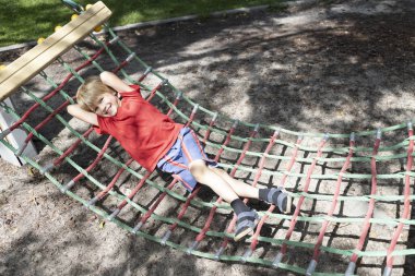 6-7 yaşlarında neşeli bir çocuk yaz günü bir parkta bir hamak ağında oturur. Çocuklar için eğlence ve eğlence. Neşeli çocukluk.