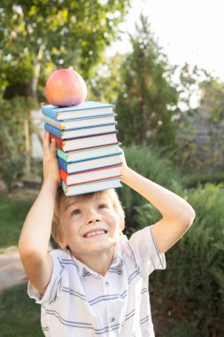 7-8 yaşlarında bir çocuk kafasında bir kaç kitap ve üzerinde bir elma tutuyor. Komik eğitim reklamı. Okuma merakı uyandırıyor. Kitap Günü. Okula geri dön.