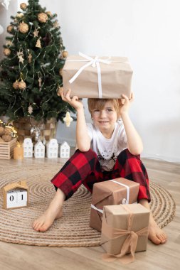 Mutlu çocuk, hediye kutularıyla bir yılbaşı ağacının önünde yerde oturuyor ve onlardan birini kafasına dayıyor. Şenlik havası. Bir Noel mucizesi bekliyorum