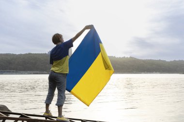 Kadın bir nehrin arka planında duruyor ve elinde bir Ukrayna sarısı-mavi bayrağı tutuyor. Ulusal özgürlük ve bağımsızlık sembolü. Ukrayna 'yı destekleyin. Gurur ve vatanseverlik. Bağımsızlık Günü