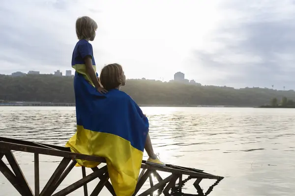 stock image small boy and woman sit with their backs turned at sunset on bank of river or lake with blue and yellow Ukrainian flag. Family, refugees, unity. Ukrainians are against war. support Ukraine