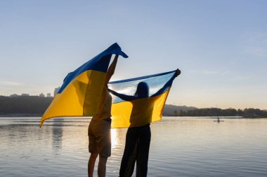Büyük Ukrayna sarı-mavi bayraklı iki kadın silueti günbatımının arka planında gökyüzü ve nehir. Ukrayna 'ya dikkat çekiyor. Anavatanı sevmek, Ukraynalı olmaktan gurur duymak.