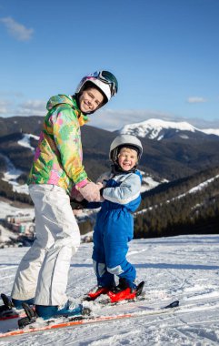 Anaokulu öğrencisi ve genç bir kız kayak pistinde el ele tutuşuyorlar. Kış sporları. Soğuk güneşli hava. Büyük ablasından küçük bir kardeşe yardım. Kayak eğitimi. Spor eğitimi