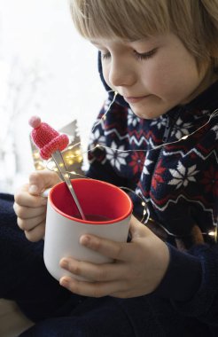 Çocuk 6 yaşında, elinde çay ya da kakao içen bir çocuğun yakın çekimi. Noel Baba şapkalı kaşık. Kışın hafta sonları ve tatillerde sıcacık bir ev. Yaşam biçimi