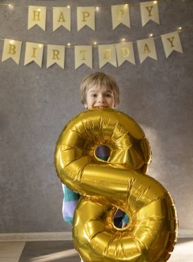 Mutlu 8 yaşındaki çocuk doğum gününü kutluyor. Doğum günü çocuğu odada duruyor ve dişlerinde sekiz numara şeklinde altın bir şenlik topu tutuyor. Doğum günü kutlamaları. Tatil, mutluluk, neşe