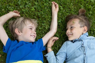 İki mutlu çocuk çimenlerin üzerinde sırt üstü yatar ve birbirleriyle neşeyle iletişim kurarlar. Bir yaz parkında dinlenen gülümseyen çocuklar. Neşe, pozitiflik, birlikte eğlenceli zamanlar. Sonsuza dek arkadaşız.