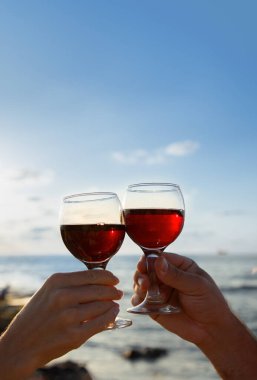 Deniz kıyısında gökyüzüne karşı cam kadehler dolusu kırmızı şarap taşıyan erkek ve kadın elleri. Doğada iki kişi için romantik bir akşam. Sevgililer Günü kutlaması