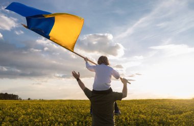 Baba ve oğul, sarı kolza tohumu tarlasında ellerinde büyük bir Ukrayna bayrağıyla omuzlarında oturuyorlar. Vatansever eğitim. Gurur, özgürlük. Ukraynalılar savaşa karşı. Aile, birlik, destek