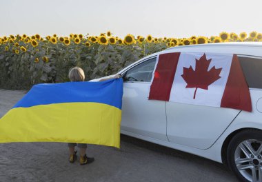 Ukrayna bayrağı taşıyan çocuk, arka planında çiçek açan Kanada bayrağı olan arabanın yanında duruyor. Ukrayna 'ya uluslararası destek ve yardım. mülteci sorunları ve göçmenlere yardım