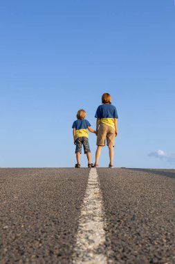 Erkek kadın, anne, mavi ve sarı tişörtlü çocuk, el ele tutuşuyorlar, asfalt yolda gökyüzüne karşı duruyorlar. Aile, mülteciler, birlik, destek, ileriye bakmak. Yaşasın Ukrayna. Ukraynalı olmanın gururu
