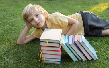 Avrupalı okul çocuğu kitap yığınlarının yanındaki çimlerin üzerinde yatıyor. Sekiz yaşındaki bir çocuk okumayı çok sever. Merhaba okul. Tatiller geldi.