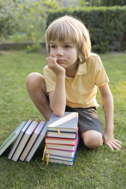 Düşünceli Avrupalı çocuk kitap yığınlarının yanındaki çimlerin üzerinde oturuyor. Sekiz yaşındaki çocuk okuma ve düşünme konusunda çok hevesli.