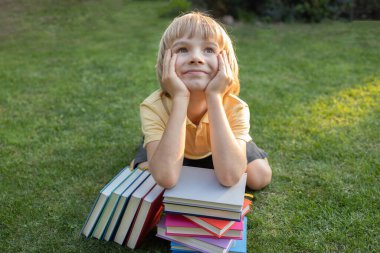 Kitap yığınının arkasında çimlerin üzerinde oturan şirin ilkokul çocuğunun portresi. Mutlu bir şekilde gülümsüyor. Kitap okumak, fantezi kurmak, ilginç bir çocukluk. Eğitim kavramı, bilgi motivasyonu