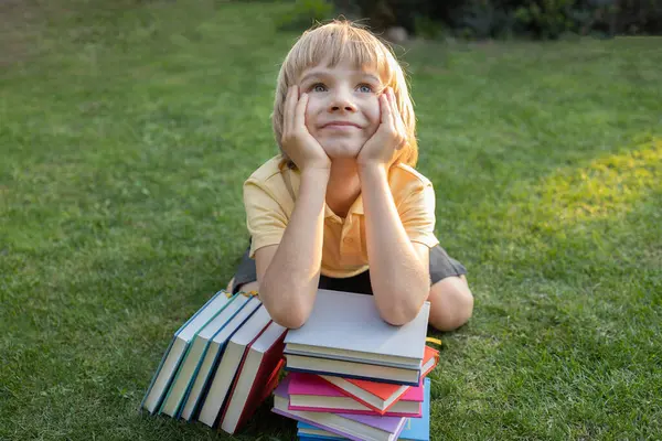 Kitap yığınının arkasında çimlerin üzerinde oturan şirin ilkokul çocuğunun portresi. Mutlu bir şekilde gülümsüyor. Kitap okumak, fantezi kurmak, ilginç bir çocukluk. Eğitim kavramı, bilgi motivasyonu