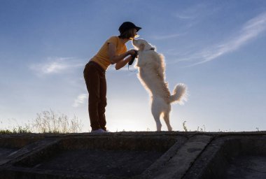 Gün batımında arka planda kadın ve köpeğin silueti gökyüzüne karşı. Doğada evcil hayvanla aktif yürüyüş. Sevgili evcil hayvandan öpücük. İletişim zevki. İnsanlar ve hayvanlar arasındaki dostluk ve sevgi
