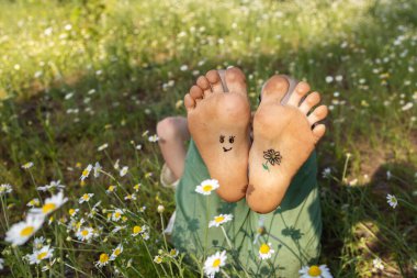 Çocuğun yalın ayakları yemyeşil çimlerin üzerinde, çiçek açan papatyaların arasında uzanıyor. Neşe, pozitif atmosfer, yaz havası, şımartma