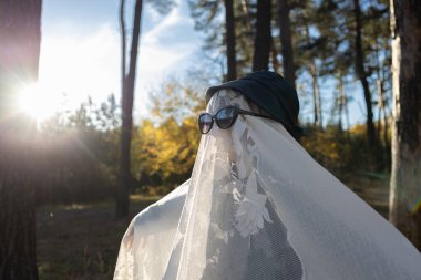 Beyaz battaniyeli ve siyah gözlüklü çocuk, güneş ışığıyla aydınlanan ormanda duruyor. Çocukların yaratıcılığı. Korkunç tatilin olumlu anları. Cadılar Bayramı 'nda iyi eğlenceler. Şeker mi şaka mı? Cadılar Bayramı konsepti