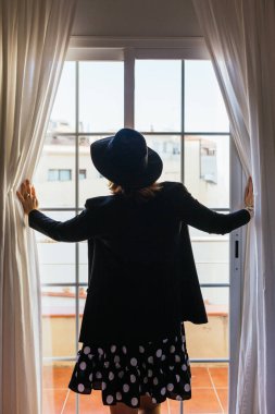 Back view of a stylish woman with hat opening the window curtains at home.