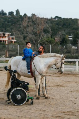 Vertical photo of a wheelchair next to a boy with cerebral palsy on top of a horse during a physiotherapy session