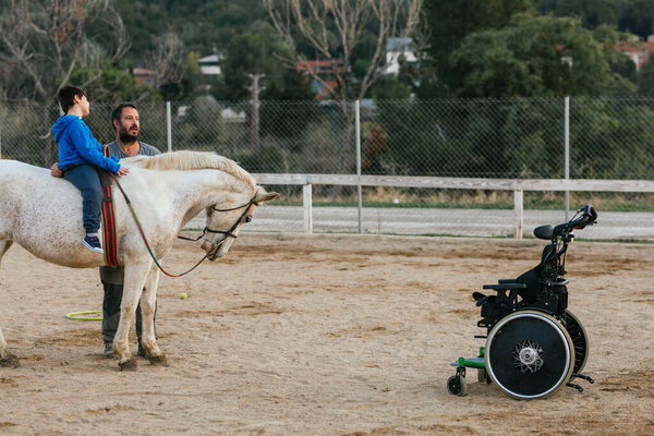 Disabled boy riding a horse next to wheelchair and his physiotherapist