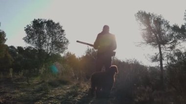 Avcı adam gün batımında köpeğiyle avlanırken av tüfeğini tutuyor. Avlanma kavramı.