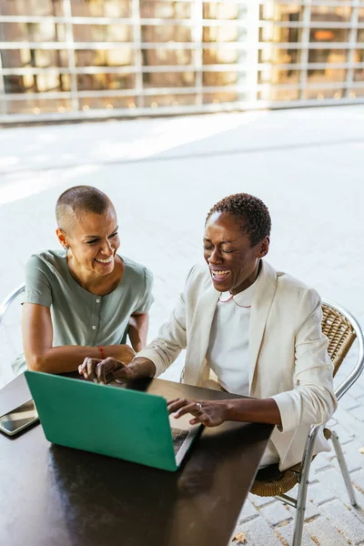 stock image Two businesswomen, one with short almost shaved hair and the other with short hair, are sitting at a street cafe, working on a laptop, and laughing. They have a mobile phone beside the laptop. They