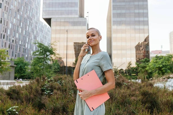 A confident and empowered woman in her 40s with short, almost shaved hair, holding a folder and answering a phone call while looking directly at the camera. She is standing in a park with business