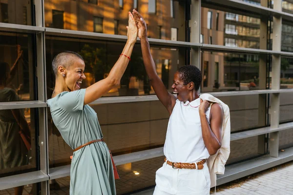 the empowering moment as two businesswomen, one Caucasian and the other African-American, enthusiastically and joyfully high-five each other in front of a business building. With their short