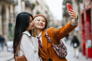Sokakta cep telefonuyla selfie çekerken gülümseyen genç lezbiyen çift. Teknoloji ve LGBT kavramı.
