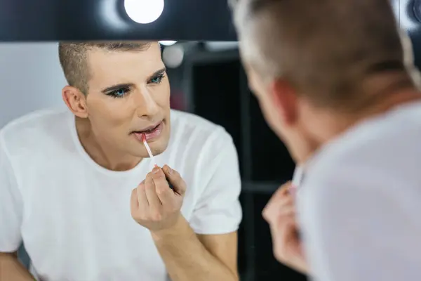 Rear view and reflection in a mirror of a gay man making up the lips facing a mirror