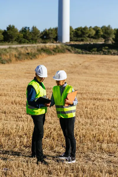 Two people in protective work clothing and reflective garments working together, discussing plans while taking notes on clipboards.