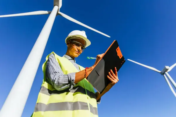 Female engineer writing on a clipboard against a wind farm, preparing a technical report for renewable energy production improvements.