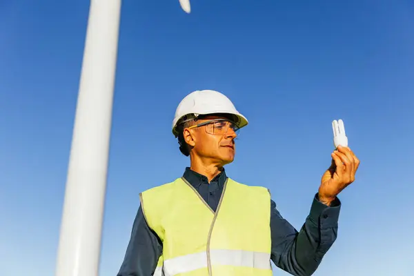Electrical engineer holding energy saving light bulb, working on windmill farm generating clean and green energy.