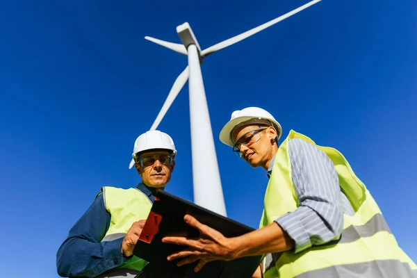 Engineer and windmill farm worker filling out paperwork, standing under a wind turbine on a sunny day.