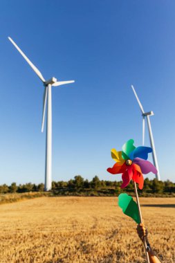 Güneşli bir günde renkli bir fırıldak tutan kişinin eli, arka planda yenilenebilir enerji üreten yel değirmenleri.