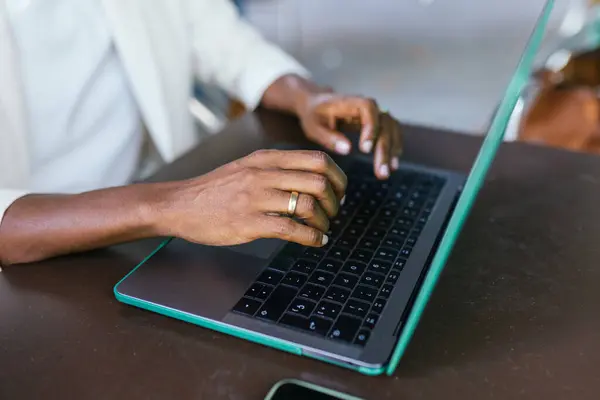 a freelancer with dark-skinned hands types on a teal laptop. The individual, dressed in a crisp white shirt, is immersed in work amidst the blurred background of a bustling coffee shop.