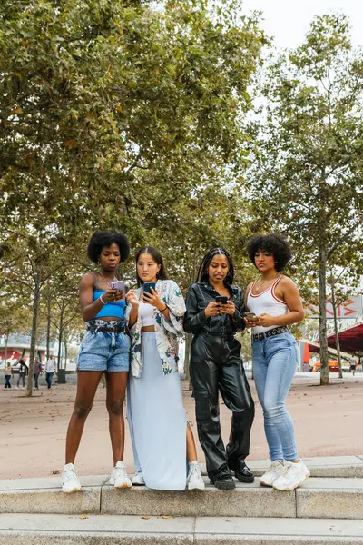 Vertical photo of diverse women and transgender person using phone standing together in a park