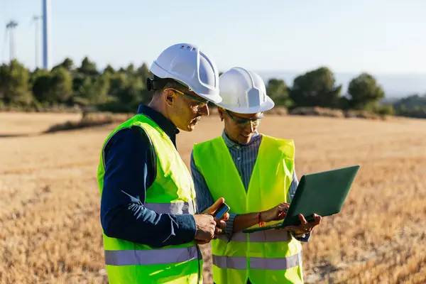 Engineers in protective work clothes and hard hats stand in a field of windmills while using digital technology.