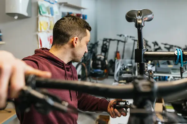 The bike mechanic holds and moves a bicycle pedal, while simultaneously testing the rear gear shift of the bike by holding the handlebar of a mountain bike..