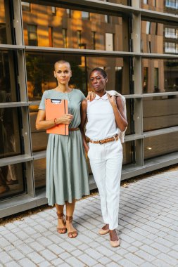 Modern ofis binasının dışında güven içinde poz veren iki profesyonel kadın. Her biri şık giyinmiş, güç ve ortaklık gösteriyor..