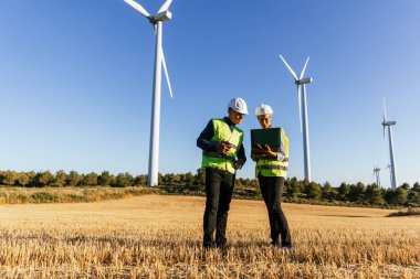 Elektrik mühendisi ve kadın sistem mühendisi yenilenebilir enerji yazılımını analiz ediyor, bir yel değirmeni çiftliğinde duruyor..