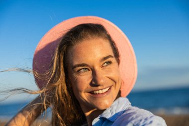 Yazlık şapka takan neşeli bir kadın ışıl ışıl gülümser, güneşli bir plaj gününde güneşlenir, sıcaklık ve mutluluğu hatırlatır.