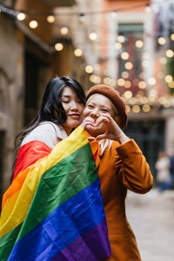 Lezbiyen çift dışarıda gökkuşağı bayrağını tutarken elleriyle kalp şekillendiriyor. LGBT kavramı.