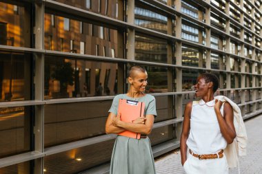 Belgeleri olan iki profesyonel kadın şirket binasının yakınında dostça sohbet ediyorlar.
