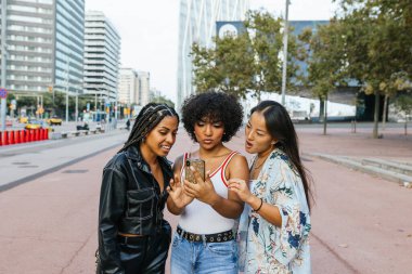 Şehrin kaldırımında birbirine yakın duran üç genç kadın. Kentsel mimariyle çevrili, gülümseyen ve paylaşılan bir anın tadını çıkaran..