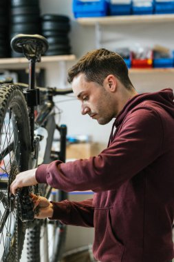 Bisiklet tamircisinin bisiklet atölyesindeki bir dağ bisikletinin arka raydan çıkarıcısının zincirini değiştirdiği dikey görüntü. Arka planda, tezgah ve lastikler görünür..
