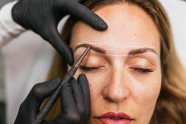Bir kadının kaş tasarımını yakından incelemek, mikroplu patinaj prosedüründen önceki şekli profesyonel güzellik araçlarıyla işaretlemek,