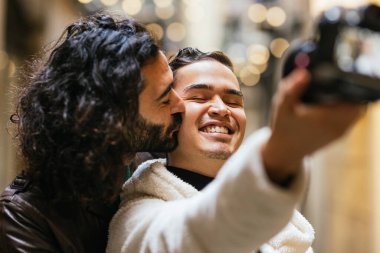 Mutlu bir anı kucaklayan ve yakalayan gülümseyen eşcinsel çift sıcak bir şehir ortamında selfie çekiyor.