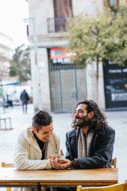 İki adam kalabalık bir şehirde açık hava masasında oturuyorlar. Çevresinden geçen şehir binaları ve yayalar ile çevrili konuşmalarla meşgul görünüyorlar..